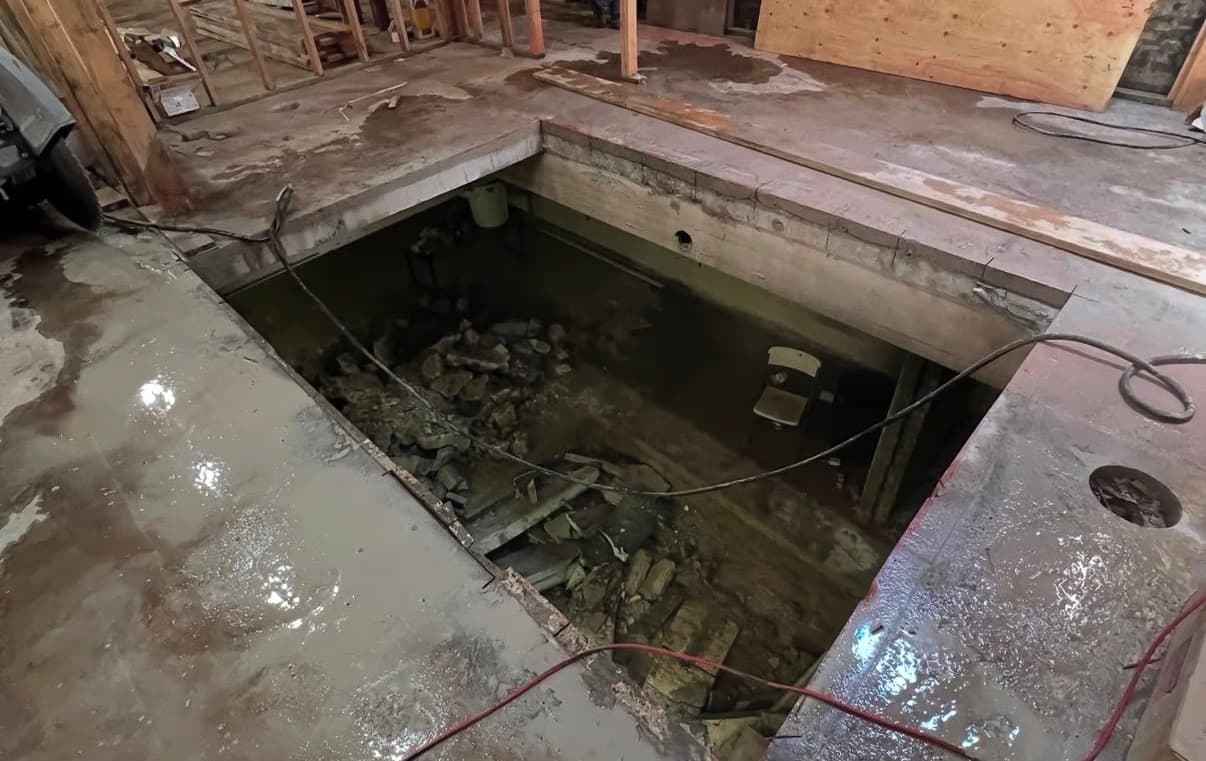 Rectangular hole in a wet concrete floor revealing a flooded basement with rocks and debris.
