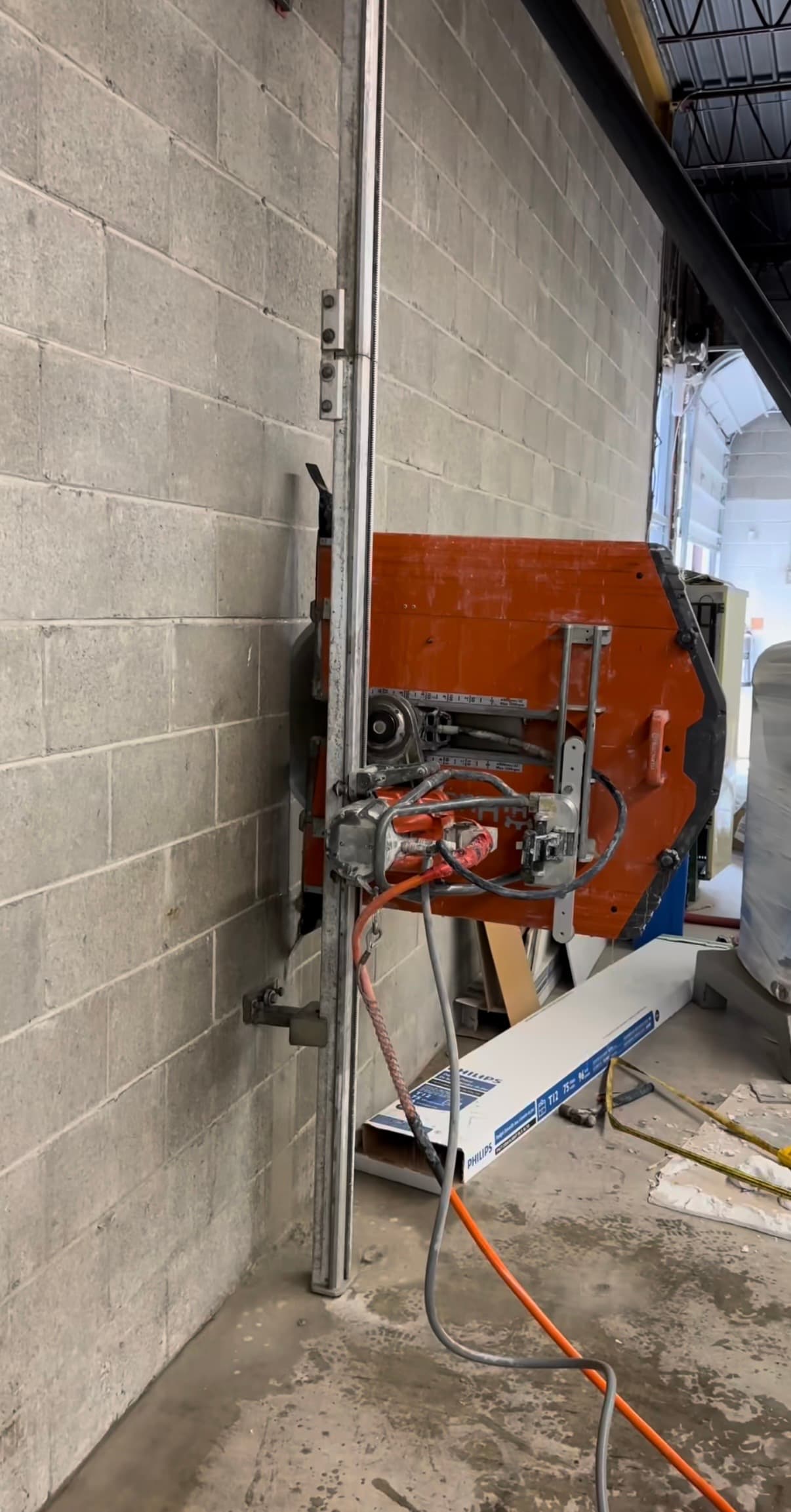 Orange wall saw mounted on a vertical track cutting into a grey concrete block wall.