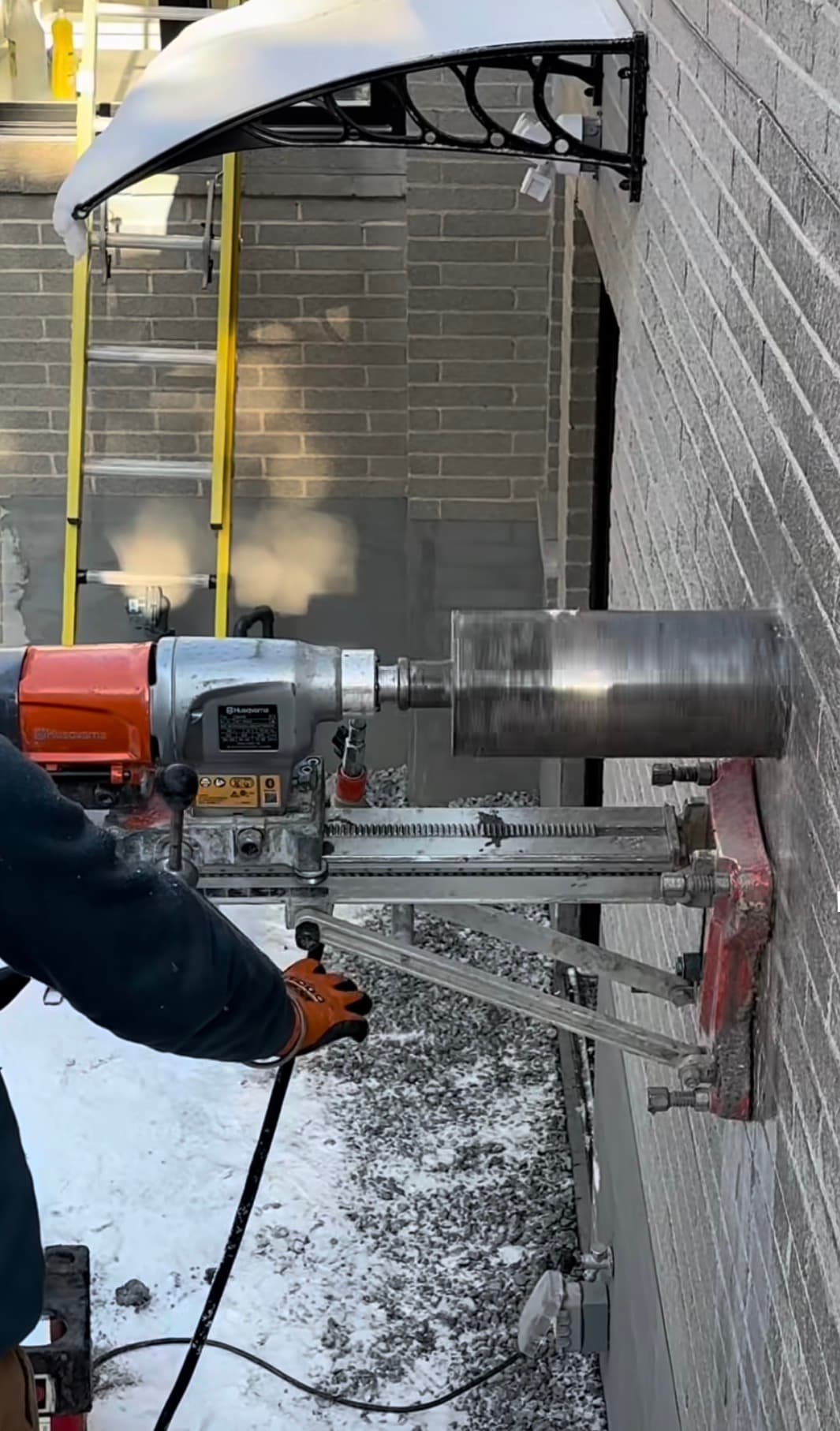 Industrial core drill machine boring a large hole into a grey brick exterior wall.