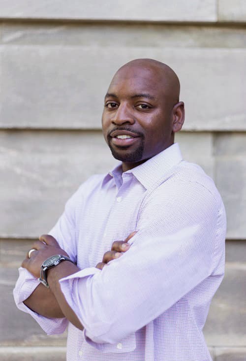 Smiling Black man with a goatee and crossed arms in a light purple shirt.